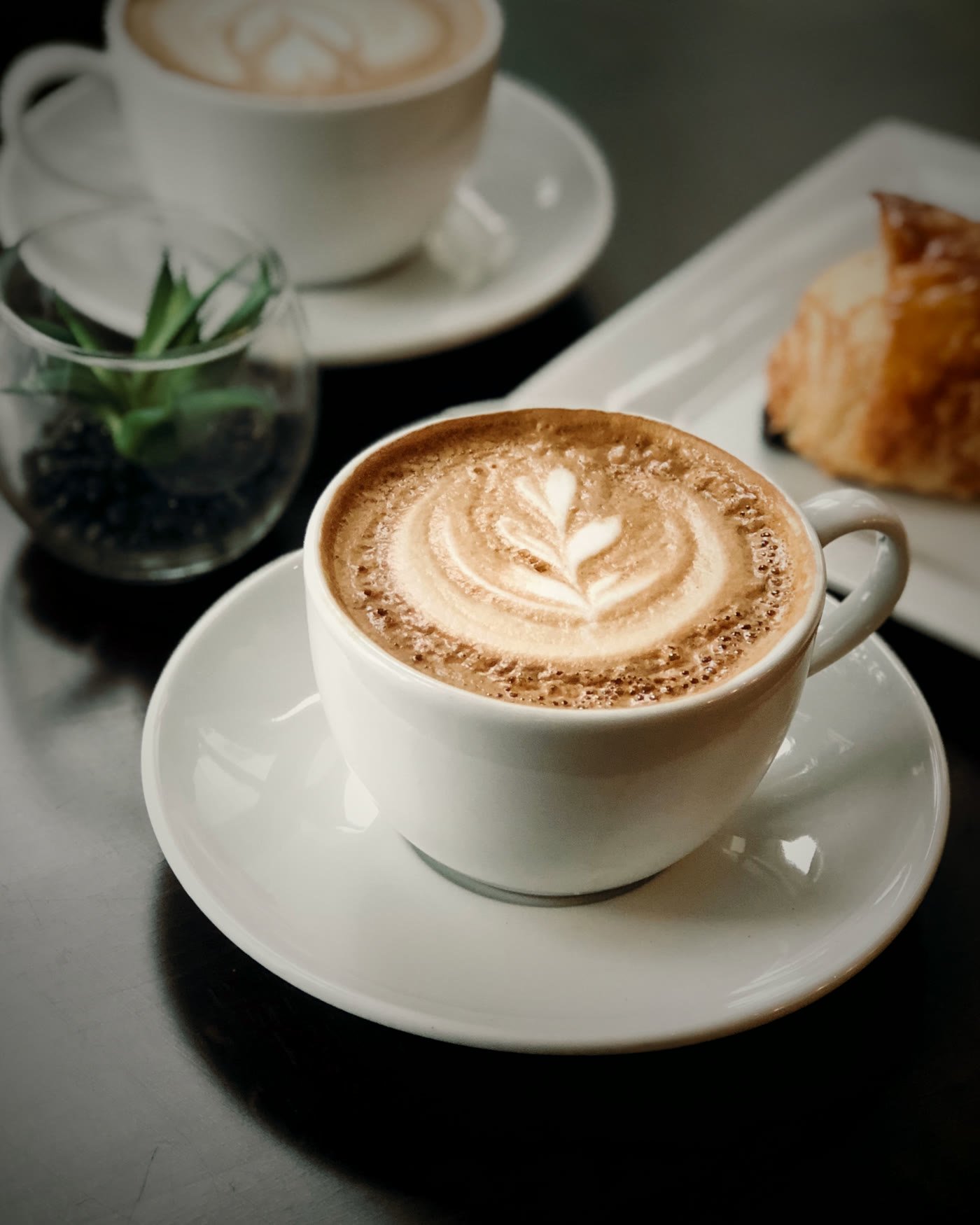 Deux tasses de cappuccino et une viennoiserie sur une table sombre.