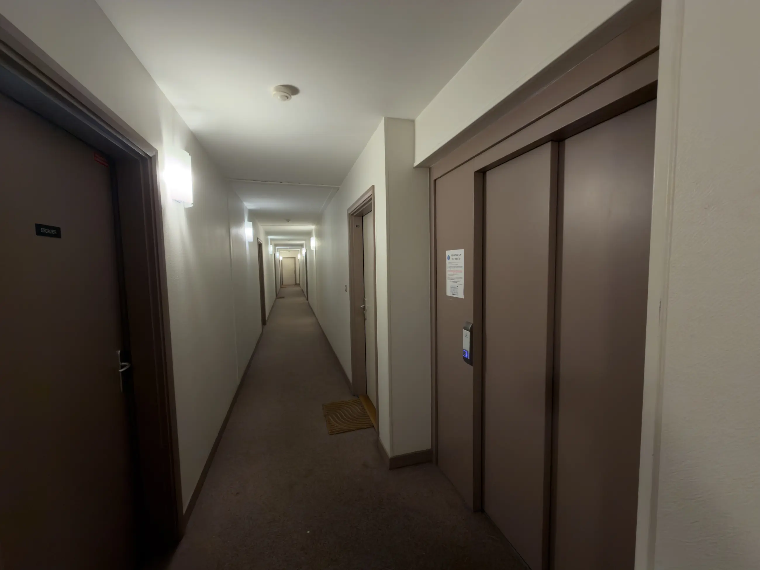 Le couloir moquetté du 2e étage, murs sans traces et appliques allumées.